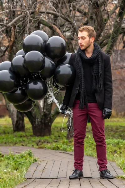 2015.11.22, Moscow, Russia. birthday is a sad holiday concept. A young blonde man wearing black scarf and coat holding black balloons and looking away.