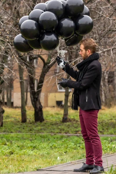 2015.11.22, Moscow, Russia. birthday is a sad holiday concept. A young blonde man wearing black scarf and coat holding black balloons and looking away.