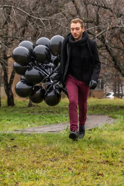 2015.11.22, Moscow, Russia. birthday is a sad holiday concept. A young blonde man wearing black scarf and coat holding black balloons running in the park.