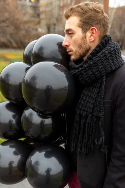 2015.11.22, Moscow, Russia. birthday is a sad holiday concept. A young blonde man wearing black scarf and coat holding black balloons and looking away.