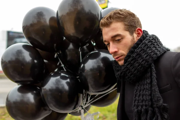 2015.11.22, Moscow, Russia. birthday is a sad holiday concept. A young blonde man wearing black scarf and coat holding black balloons and looking away.