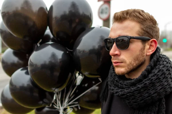 2015.11.22, Moscow, Russia. birthday is a sad holiday concept. A young blonde man wearing sunglasses and black scarf holding black balloons, side view.