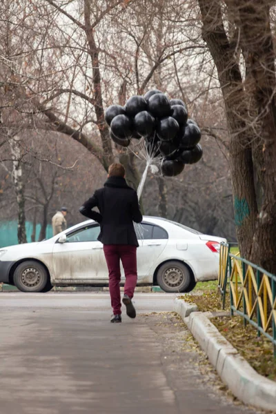 2015.11.22, Moscow, Russia. birthday is a sad holiday concept. A young blonde man wearing black scarf and coat holding black balloons going down street, back side view.