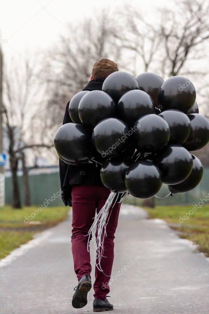 2015.11.22, Moscow, Russia. birthday is a sad holiday concept. A young blonde man wearing black scarf and coat holding black balloons going down street, back side view.