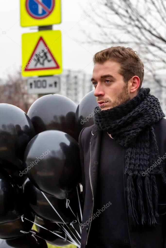 2015.11.22, Moscow, Russia. birthday is a sad holiday concept. A young man wearing black scarf and coat holding black balloons on background of urban scene, blur and grain effect.