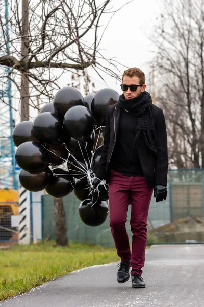 2015.11.22, Moscow, Russia. birthday is a sad holiday concept. A young blonde man wearing black scarf and coat holding black balloons going down street, front view.