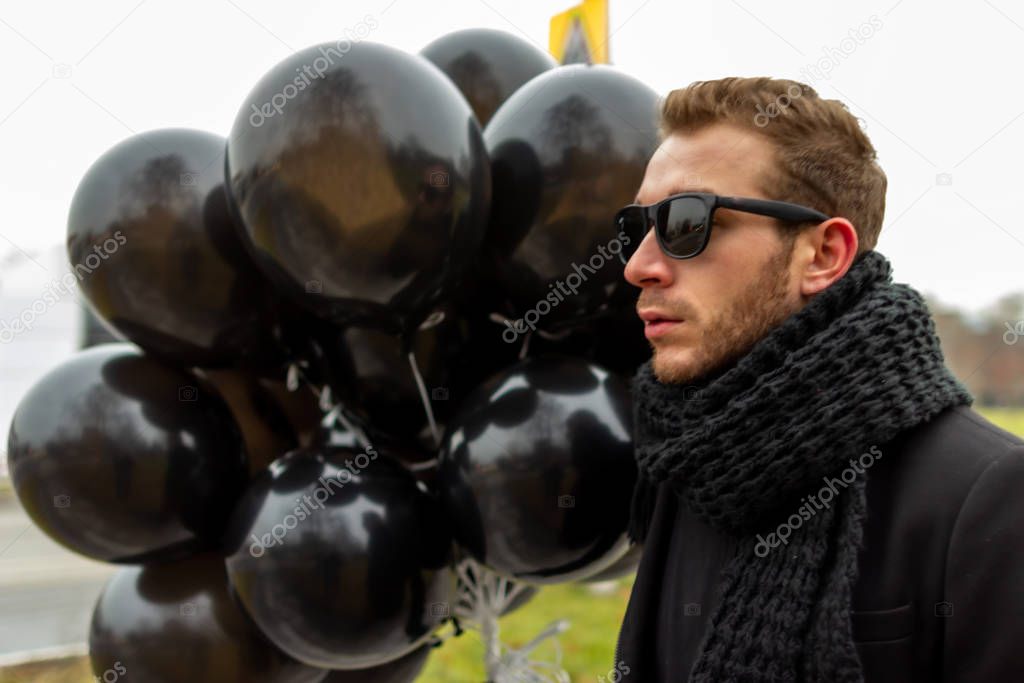 2015.11.22, Moscow, Russia. birthday is a sad holiday concept. A young blonde man wearing sunglasses and black scarf holding black balloons, side view.