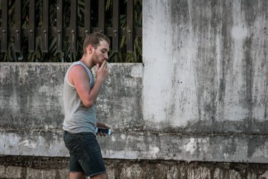 Gri kolsuz tişörtlü genç adam ve kot pantolonlar beton bir çitin arkasında sigara içiyor.