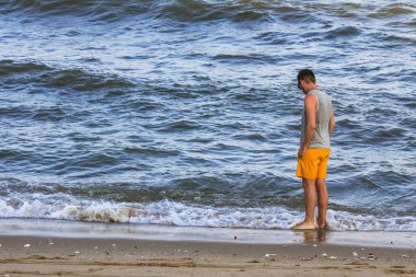 Sarı şortlu ve gri tişörtlü genç adam kıyıda duruyor ve okyanusa bakıyor.