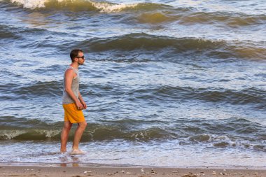 Sarı şort ve gri tişört giyen genç adam kıyıda yürüyor ve okyanusa bakıyor.