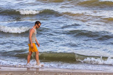 Sarı şort ve gri tişört giyen genç adam kıyıda yürüyor ve okyanusa bakıyor.