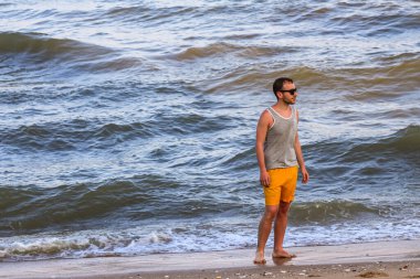 Sarı şortlu ve gri tişörtlü genç adam kıyıda duruyor ve okyanusa bakıyor.