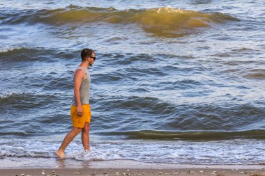 Sarı şort ve gri tişört giyen genç adam kıyıda yürüyor ve okyanusa bakıyor.
