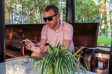 Genç, yakışıklı, sakallı, pembe polo ve siyah güneş gözlüğü takan adam lobide oturmuş telefona bakıyor.