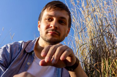 Kot pantolon, beyaz tişört ve mavi tişört giyen genç çekici bir adam mavi gökyüzüne karşı mısır gevreği tarlasında uzanıyor ve kameraya bakıyor.