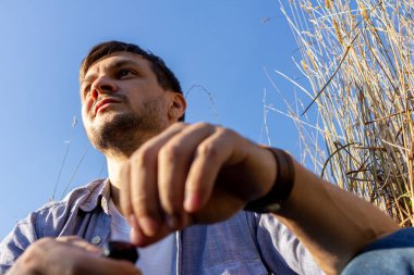 Kot pantolon, beyaz tişört ve mavi tişört giyen genç çekici bir adam mavi gökyüzüne karşı mısır gevreği tarlasında yatar ve sola bakar.