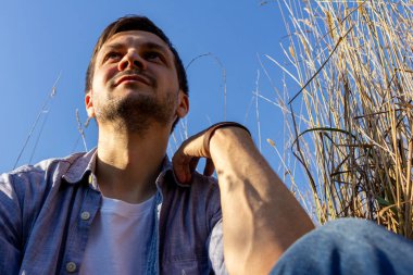 Kot pantolon, beyaz tişört ve mavi gömlek giyen genç çekici bir adam mısır gevreği tarlasında yatar ve yukarı bakar..