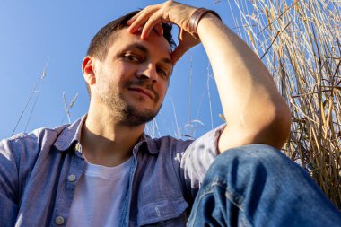 Kot pantolon, beyaz tişört ve mavi tişört giyen genç çekici bir adam mavi gökyüzüne karşı mısır gevreği tarlasında uzanıyor ve kameraya bakıyor.