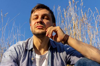 Kot pantolon, beyaz tişört ve mavi tişört giyen genç çekici bir adam mavi gökyüzüne karşı mısır gevreği tarlasında uzanıyor ve kameraya bakıyor.