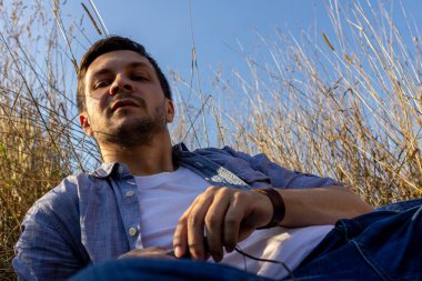 Kot pantolon, beyaz tişört ve mavi tişört giyen genç çekici bir adam mavi gökyüzüne karşı mısır gevreği tarlasında uzanıyor ve kameraya bakıyor.