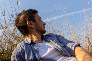 Kot pantolon, beyaz tişört ve mavi tişört giyen genç çekici bir adam mavi gökyüzüne karşı mısır gevreği tarlasında yatar ve sağa bakar.