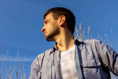 Kot pantolon, beyaz tişört ve mavi tişört giyen genç çekici bir adam mavi gökyüzüne karşı mısır gevreği tarlasında yatar ve sola bakar.