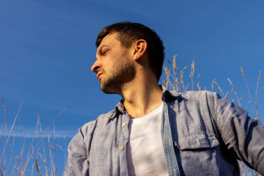 Kot pantolon, beyaz tişört ve mavi tişört giyen genç çekici bir adam mavi gökyüzüne karşı mısır gevreği tarlasında yatar ve sola bakar.