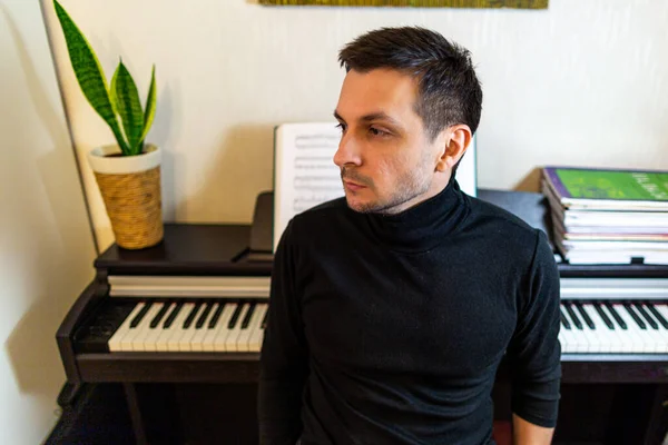 A dark-haired tanned young guy wearing a black turtleneck sitting with his back to the digital piano, looking to the left.