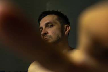 Genç, çekici, üstsüz adam karanlık banyoda oturuyor ve kendi parmaklarından bakıyor. .