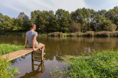 Açık renk şort ve tişört giyen genç bir adam nehir kıyısındaki ahşap bir yapının üzerinde oturuyor ve nehre, arka ve yan manzaraya bakıyor. Rahatlatıcı hafta sonu.