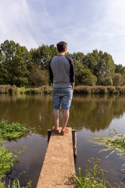 Kot şortlu ve uzun kollu genç bir adam nehir kenarındaki ahşap bir yapının üzerinde duruyor, elleri ceplerinde ve nehre bakıyor, arkadan ve yandan bakıyor..