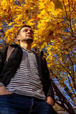Sonbahar ağacının altında duran siyah ceketli kısa saçlı genç bir adam. Düşen parkta meditasyon.