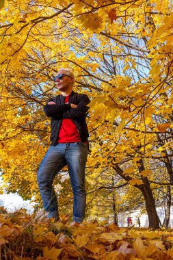 Sonbahar parkında kollarını kavuşturmuş siyah ceket giyen kısa saçlı genç bir adam. Düşen parkta meditasyon.