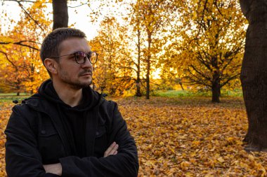 Genç, kısa saçlı, gözlüklü ve siyah ceketli bir adam sonbahar parkında kollarını kavuşturmuş duruyor. Düşen parkta meditasyon.