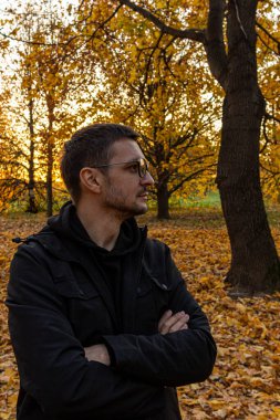 Genç, kısa saçlı, gözlüklü ve siyah ceketli bir adam sonbahar parkında kollarını kavuşturmuş duruyor. Düşen parkta meditasyon.