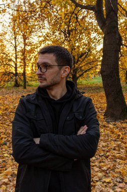 Genç, kısa saçlı, gözlüklü ve siyah ceketli bir adam sonbahar parkında kollarını kavuşturmuş duruyor. Düşen parkta meditasyon.