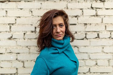 Mavi örgü elbise giyen oldukça uzun saçlı genç bir kadın beyaz tuğla bir duvara bakıyor ve kameraya bakıyor..