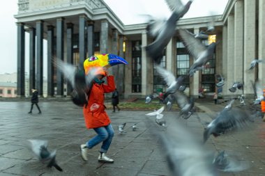 Rus devlet kütüphanesinin yanındaki meydandaki güvercin sürüsünün arasında koşan kırmızı ceketli ve maskeli genç bir kadın..