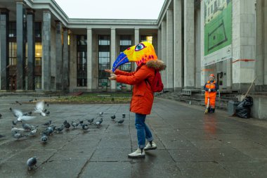 Kırmızı ceket ve maske takan genç bir kadın, Rus devlet kütüphanesinin yakınındaki meydanda bir güvercin sürüsünü besliyor..