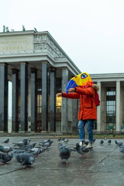 Kırmızı ceket ve maske takan genç bir kadın, Rus devlet kütüphanesinin yakınındaki meydanda bir güvercin sürüsünü besliyor..
