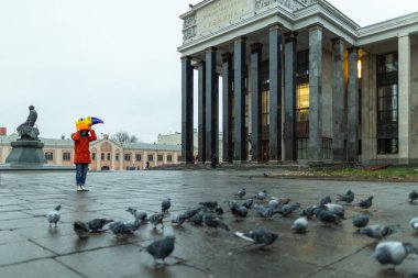 Rus devlet kütüphanesinin yanındaki meydandaki güvercin sürüsünün arasında koşan kırmızı ceketli ve maskeli genç bir kadın..