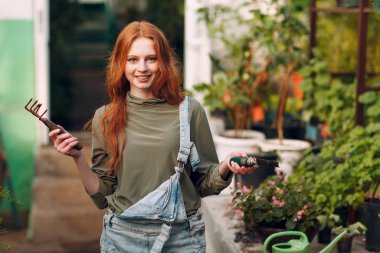 Ev işleri konsepti. Kot tulumlu, tırmık ve kürekle bitki eken genç kızıl kadın. Bahar bahçesi bitkisi.