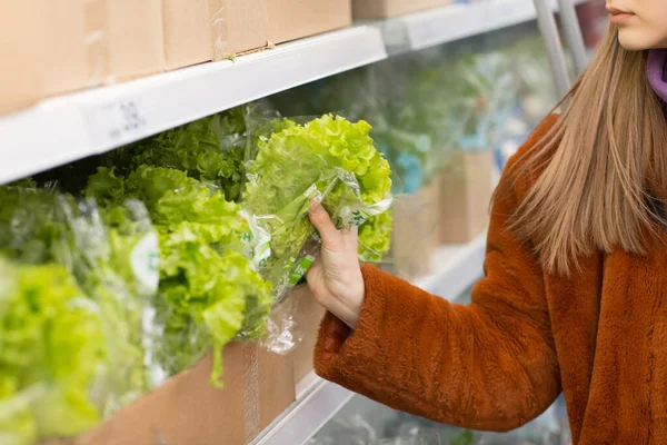 Young woman with a grocery basket chooses products in the vegetable department. Green salad.