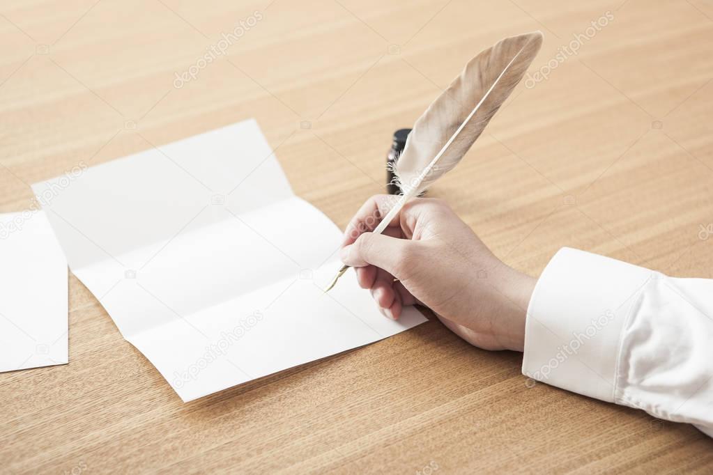 man dipping a feather pen into an ink pot to write a letter on daylight wood desk.
