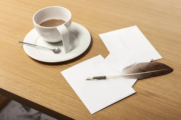 A coffee cup, feather quill pen, letter, envelope on the office desk(table) behind white blind.