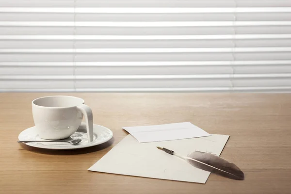 A coffee cup, feather quill pen, letter, envelope on the office desk(table) behind white blind.