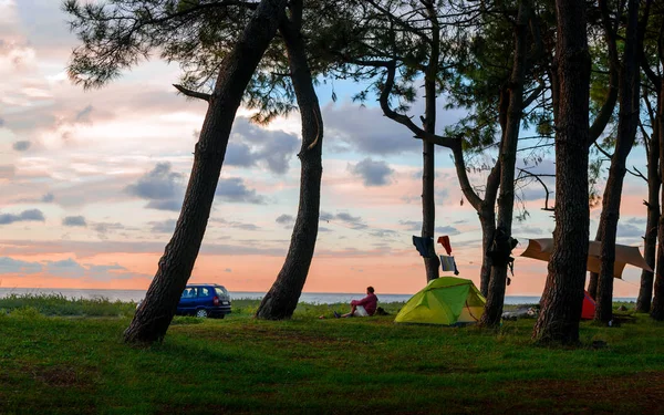 Hombre sentado al atardecer en la playa cerca de tiendas de campaña bajo los árboles —  Fotos de Stock