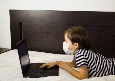 Karantinadaki bir çocuk çevrimiçi eğitim yapıyor. Evde ödev yapıyorum. Laptop yatağın üstünde. Tıbbi maske