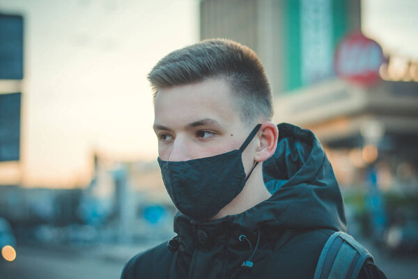 Closeup portrait of young guy in medical mask. Pandemic Coronavirus. Quarantine Covid 19. Student in a protective mask. Gloomy young man in black protective mask and black clothing in the city.