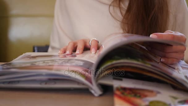 Joven hermosa chica hojeando a través de una revista en un café, primer plano, 4k — Vídeo de stock